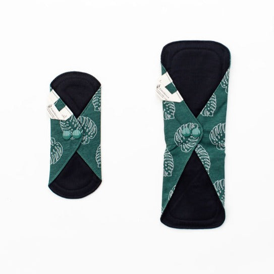 two feminine pads in liner and daytime size. The pads have a dark green with lighter green leaf-like motifs